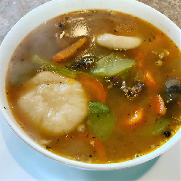 Steaming bowl of Jamaican soup with dumplings, carrots, and green vegetables.