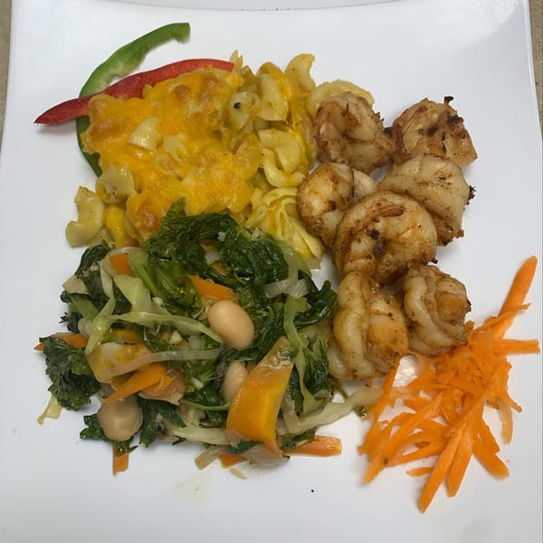 A Jamaican meal with seasoned shrimp, cheesy macaroni, sautéed greens, and shredded carrots on a white plate.